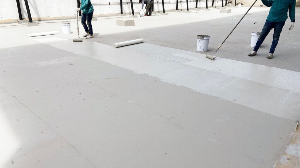 Two roofing contractors are using paint rollers to apply a white roof coating to the surface of a flat roof in Tucson