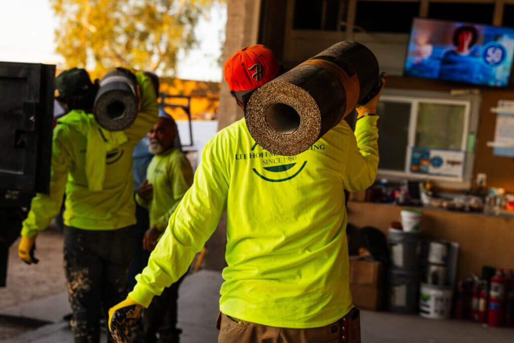 Two roofing contractors are carrying rolls of new roof underlayment on their shoulders out of a garage at a jobsite