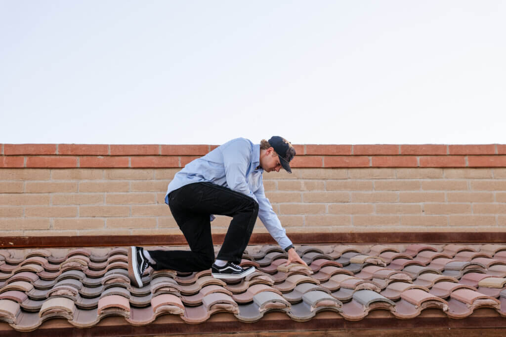 A roofing contractor is kneeling on a tile roof on a home in Tucson, inspecting the tiles for signs of damage