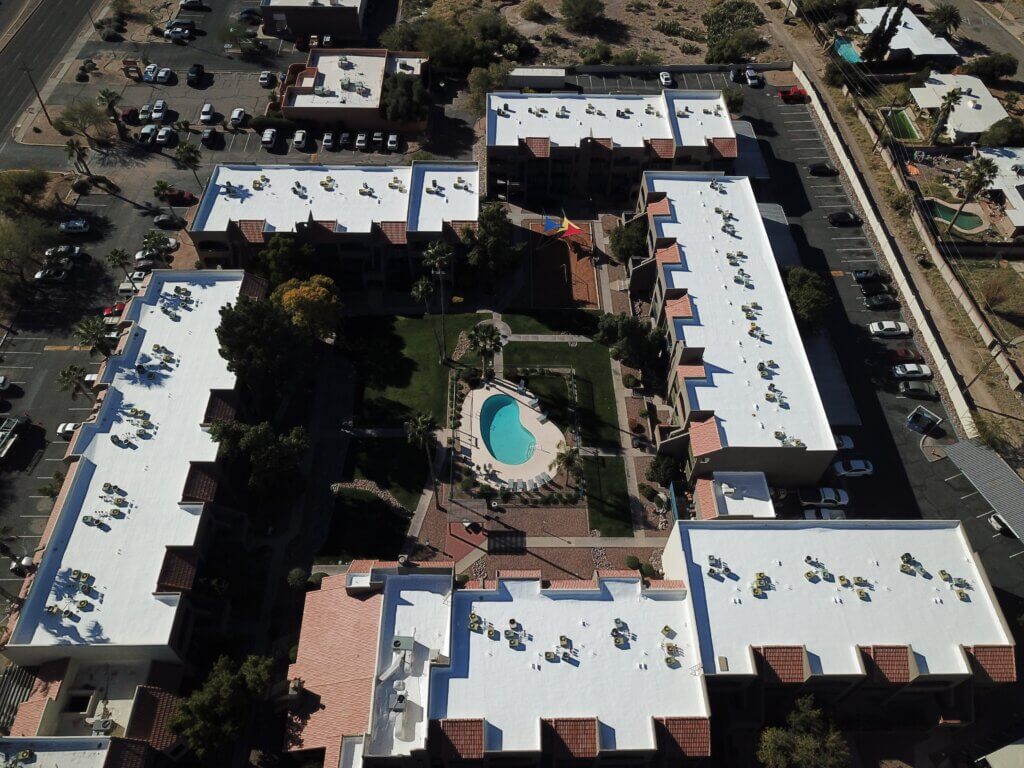 An apartment complex of 5 buildings, all with new white roof coatings, are in a rectangle around a park and swimming pool