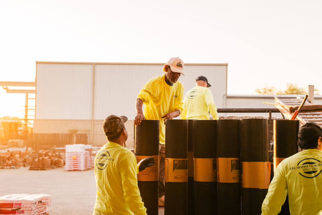 Four roofing contractors are loading large rolls of roof underlayment into the bed of a pickup truck